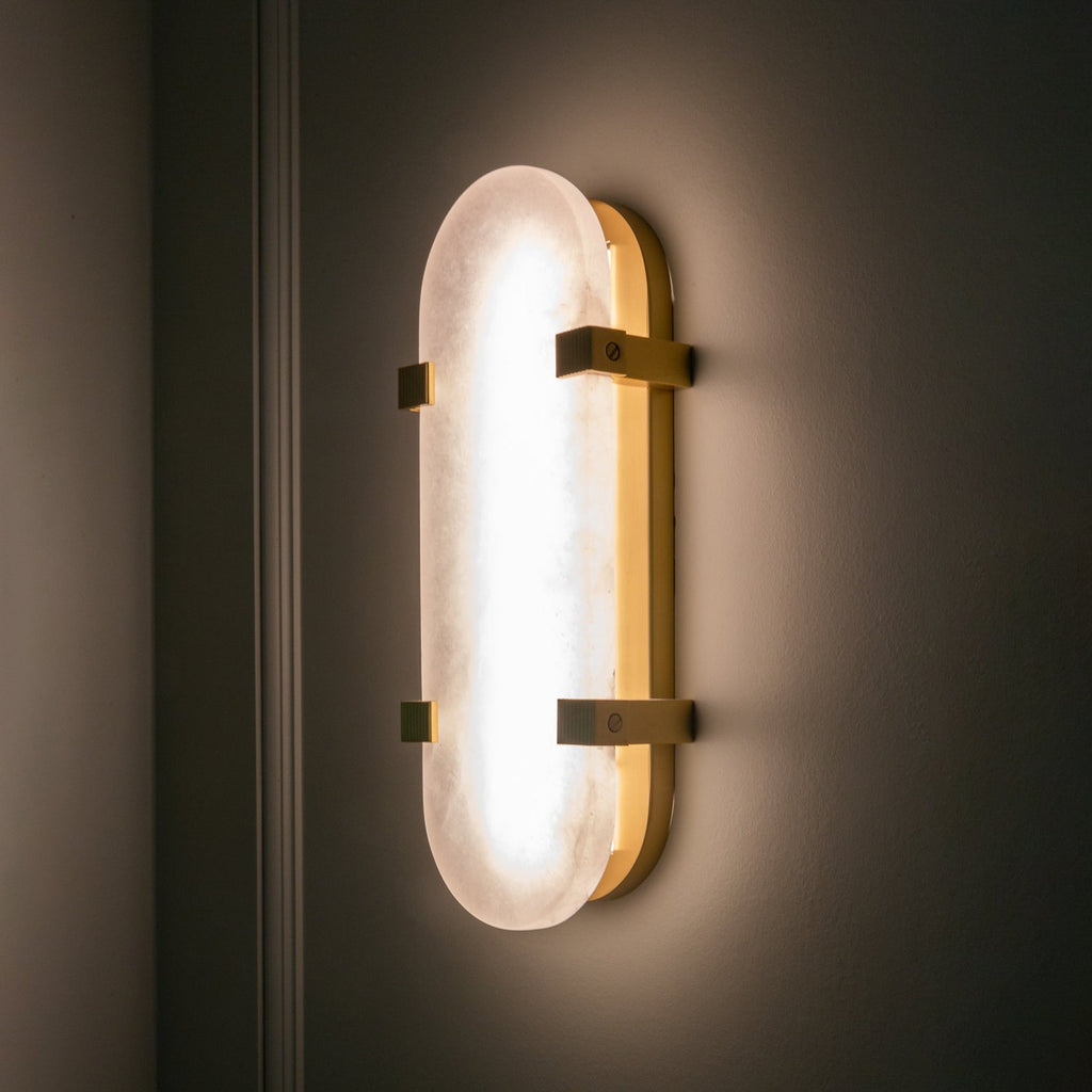 Lit Clérachic wall light glowing evenly on a dark wall, its alabaster shade framed by brass clips and a slim brass edge.