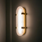 Lit Clérachic wall light glowing evenly on a dark wall, its alabaster shade framed by brass clips and a slim brass edge.