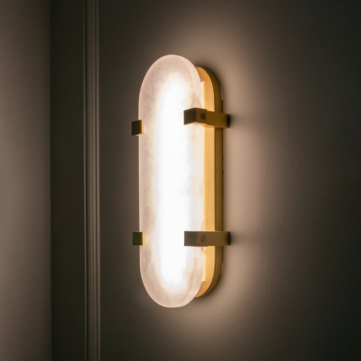 Lit Clérachic wall light glowing evenly on a dark wall, its alabaster shade framed by brass clips and a slim brass edge.
