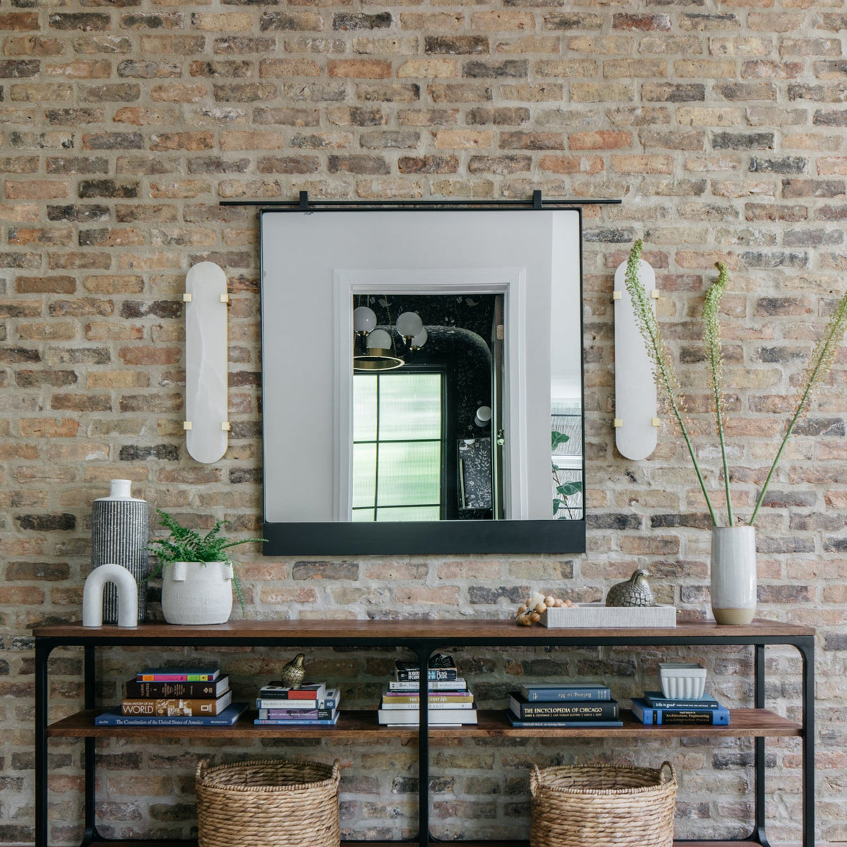Two Clérachic wall lights flank a black-framed mirror on an exposed brick wall above a dark console with books and baskets.