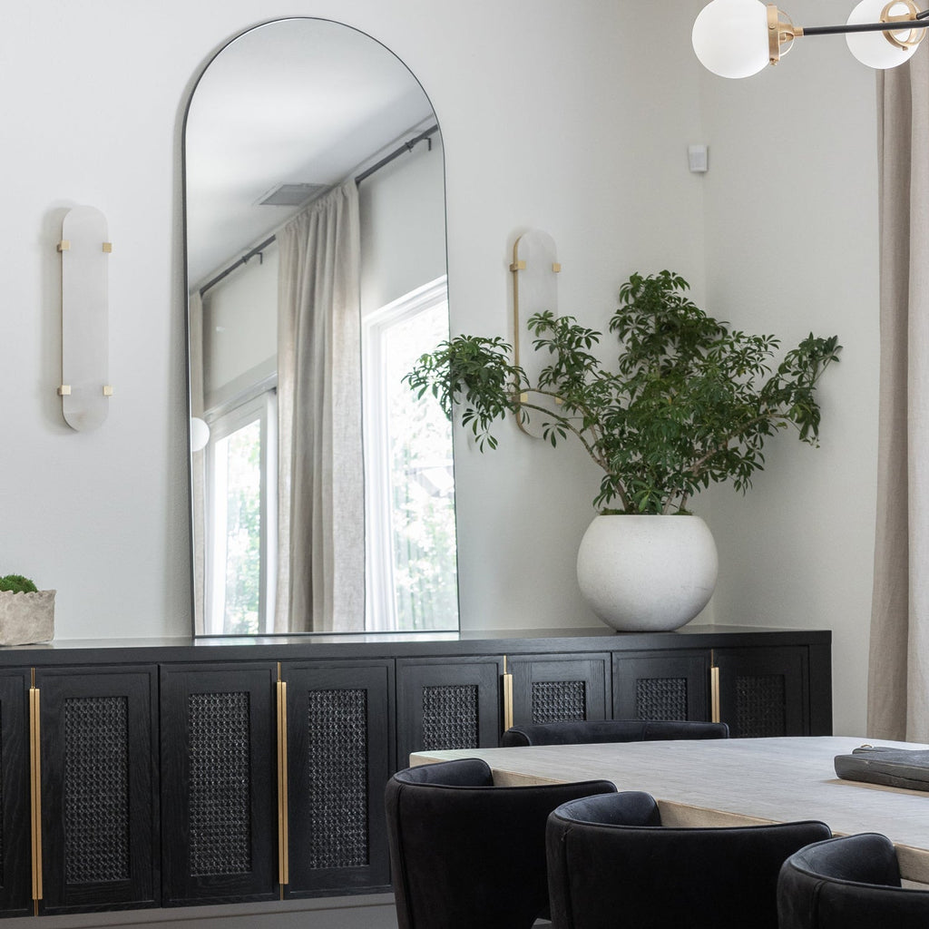 A pair of Clérachic wall lights on a pale wall beside a tall arched mirror, with a black sideboard and a leafy plant below.