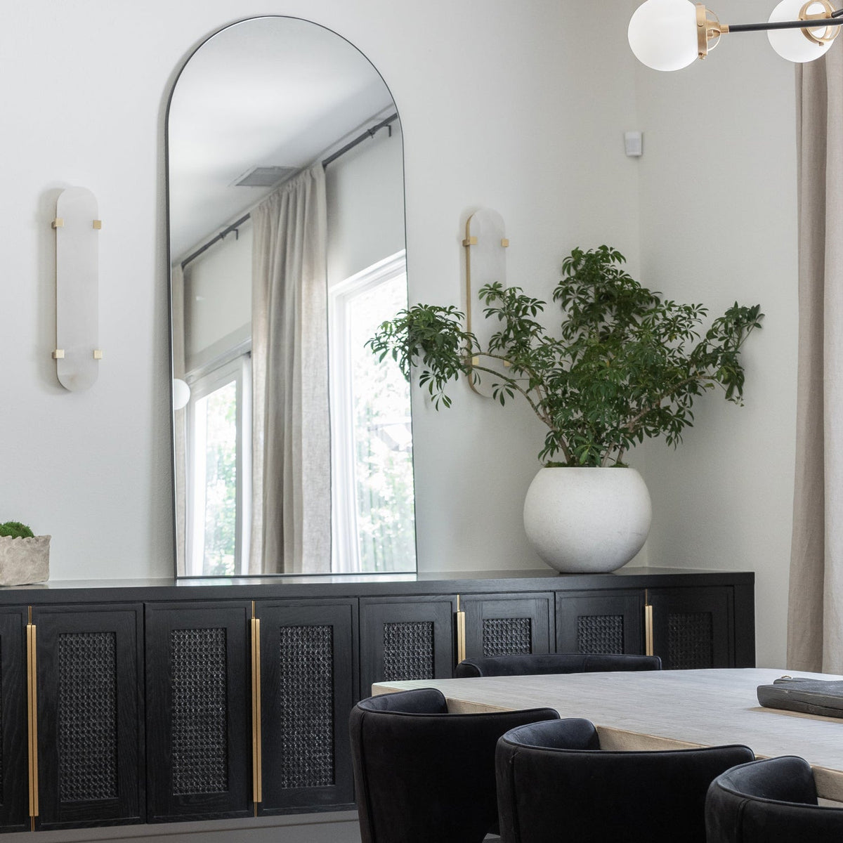 A pair of Clérachic wall lights on a pale wall beside a tall arched mirror, with a black sideboard and a leafy plant below.