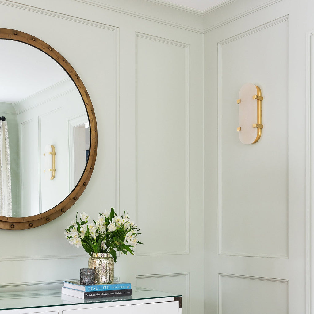 Unlit Clérachic wall light on a panelled sage-green wall, next to a round mirror and a vase of white flowers on a console.