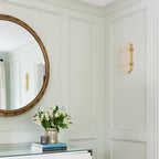 Unlit Clérachic wall light on a panelled sage-green wall, next to a round mirror and a vase of white flowers on a console.