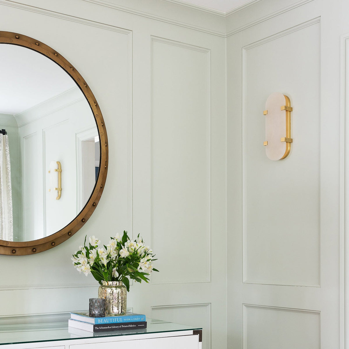 Unlit Clérachic wall light on a panelled sage-green wall, next to a round mirror and a vase of white flowers on a console.