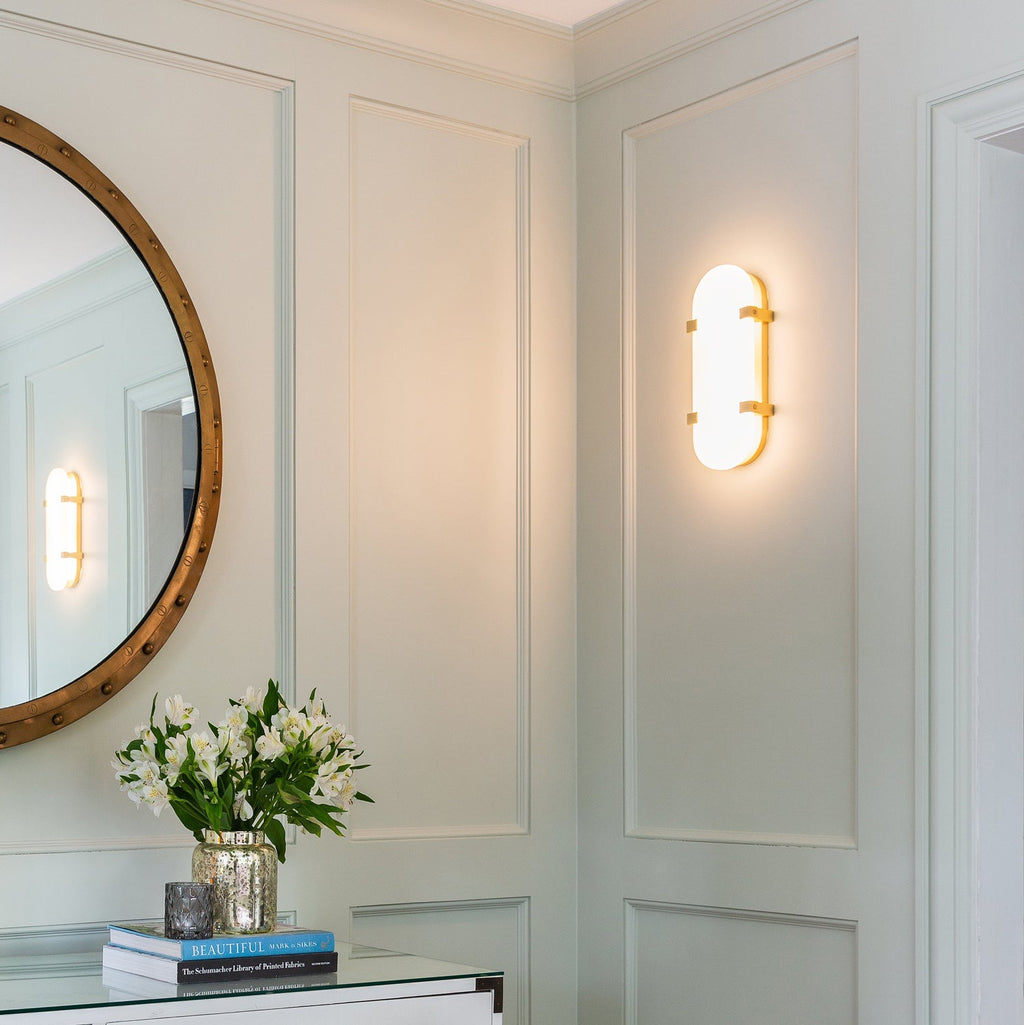 Lit Clérachic wall light giving a warm glow on a panelled green wall, with a round brass-edged mirror and white flowers below.