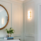 Lit Clérachic wall light giving a warm glow on a panelled green wall, with a round brass-edged mirror and white flowers below.