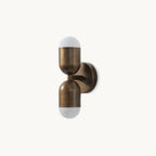 A Clérachic wall light in antique brass with two capsule shades, one above the other, on a plain white backdrop.