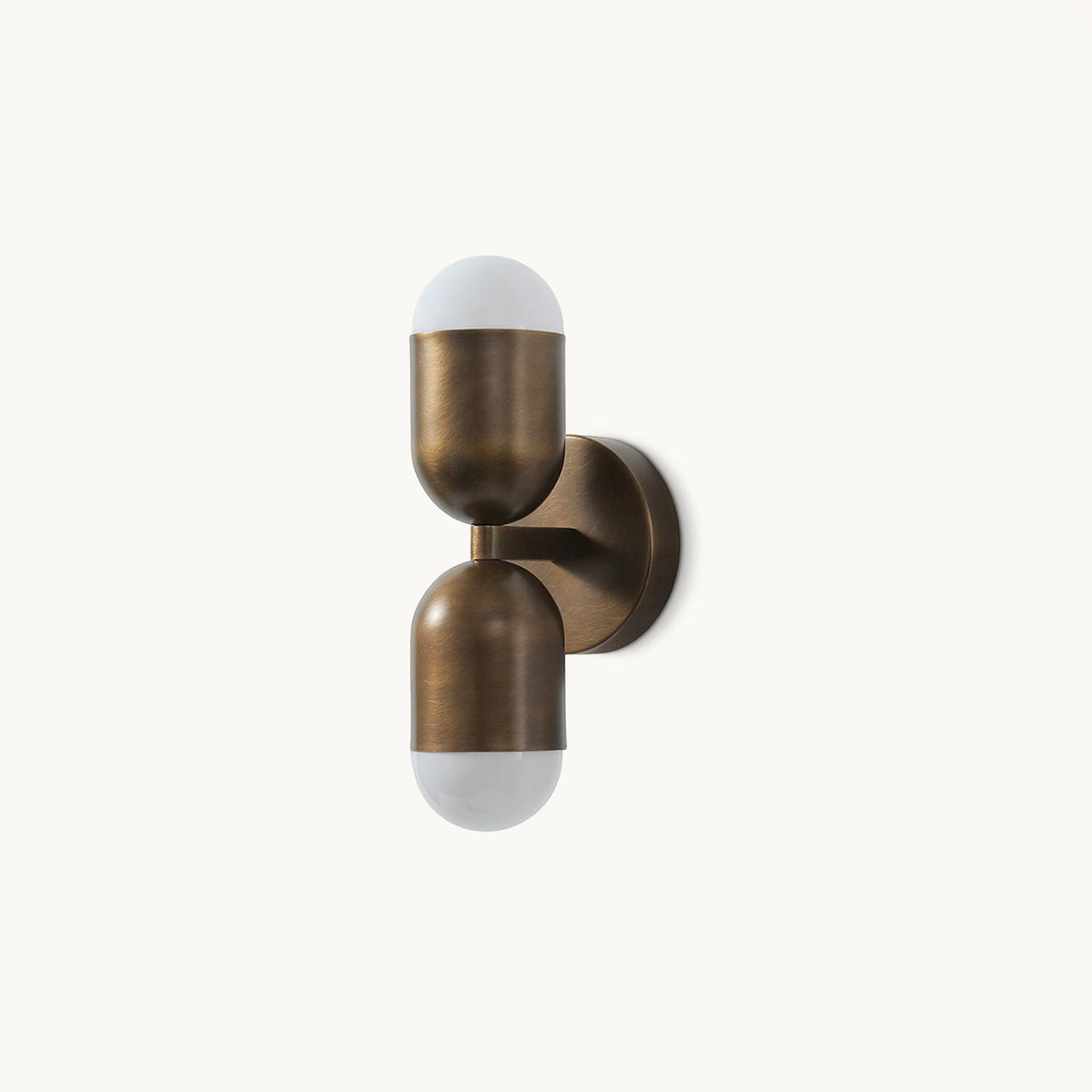 A Clérachic wall light in antique brass with two capsule shades, one above the other, on a plain white backdrop.