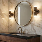 Two antique brass Clérachic wall lights on either side of a round brass-framed mirror above a marble-tiled bathroom vanity.