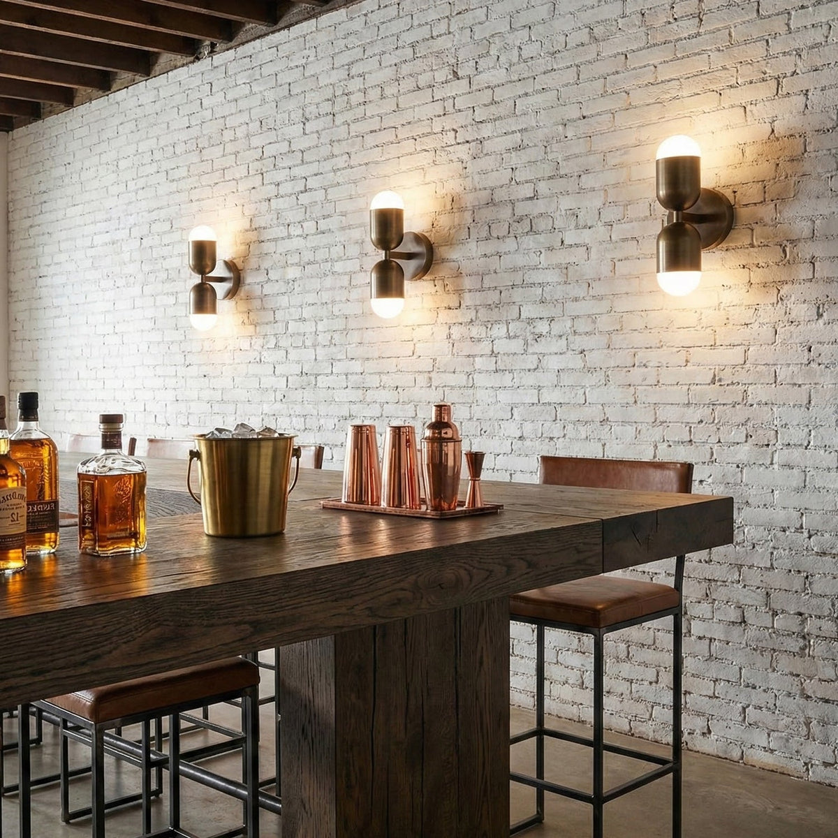 Three antique brass Clérachic wall lights glowing along a white brick wall above a dark wood bar table.