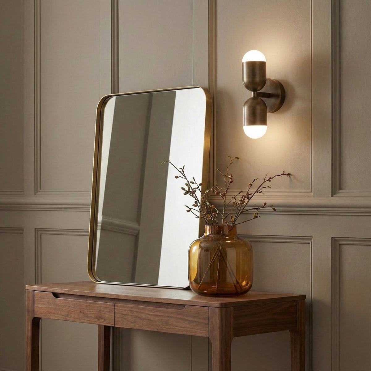 An antique brass Clérachic wall light on a panelled beige wall, above a walnut console with a mirror and an amber vase.