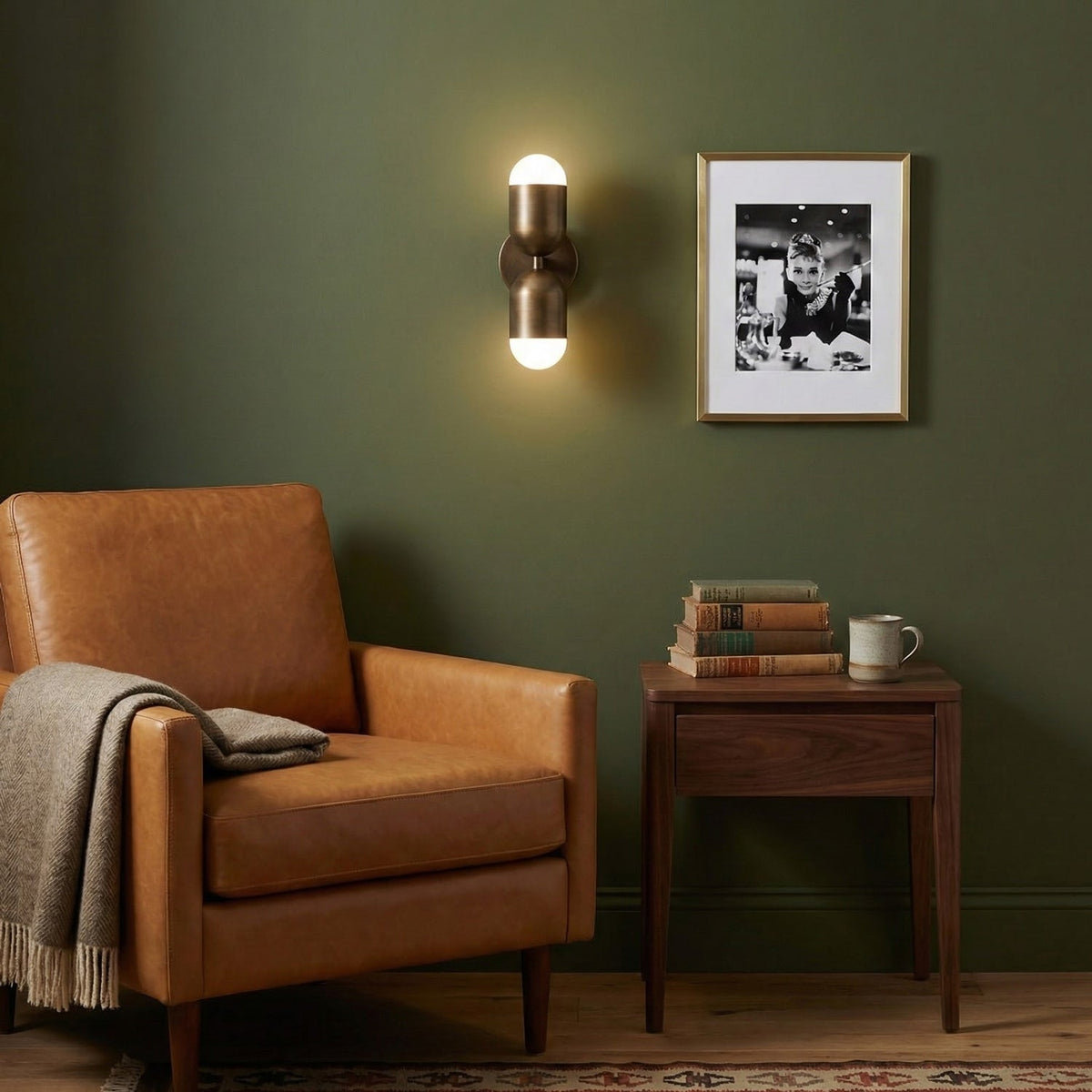 Antique brass Clérachic wall light on an olive-green wall, above a tan leather armchair and a small side table of books.