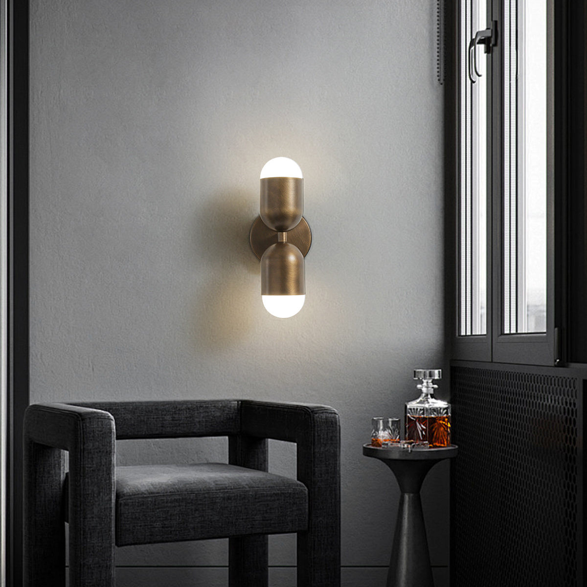Antique brass Clérachic wall light on a grey wall in a window corner, above a dark armchair and a small drinks table.