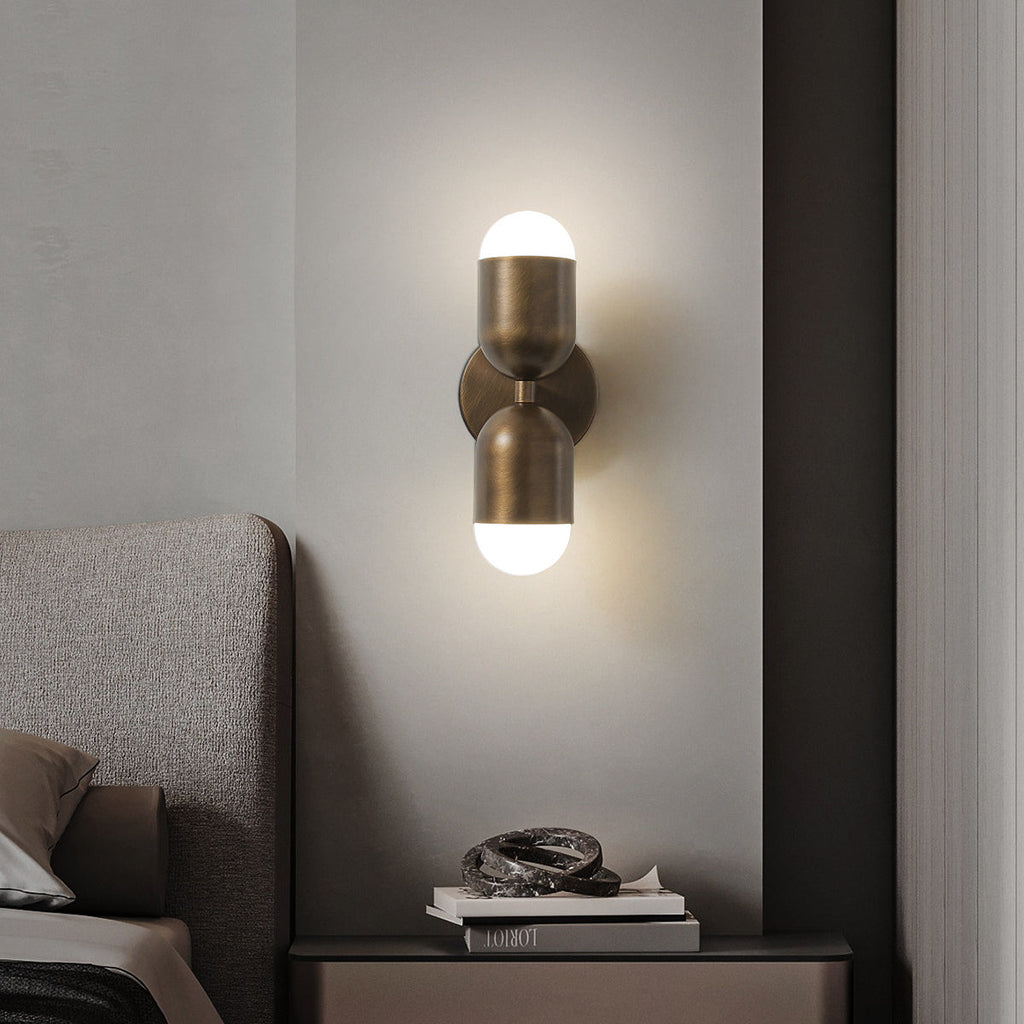 A Clérachic wall light with two brass capsule shades, lit top and bottom, on a pale wall above a bedside table.
