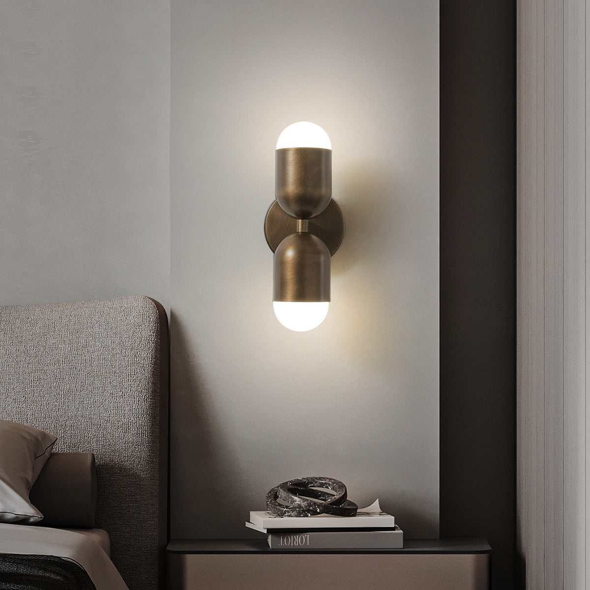 A Clérachic wall light with two brass capsule shades, lit top and bottom, on a pale wall above a bedside table.