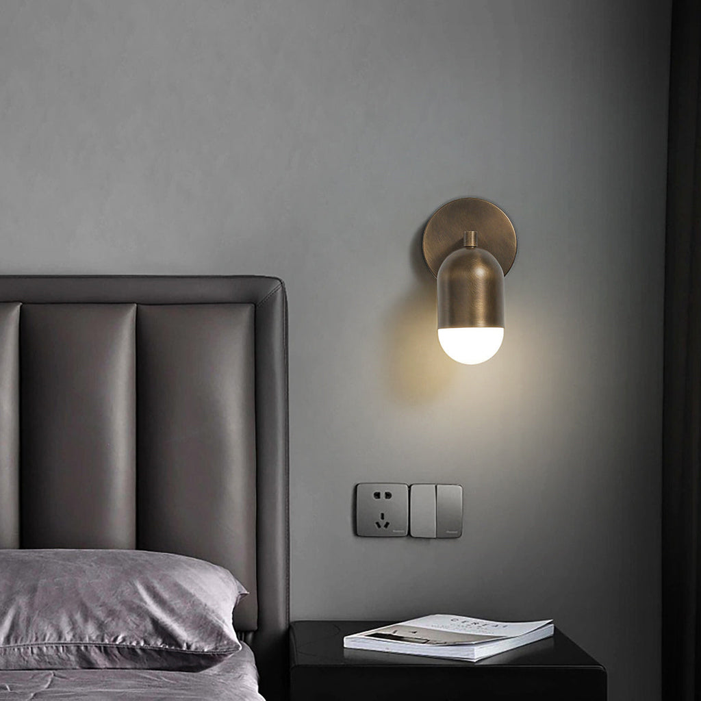 Single-capsule antique brass Clérachic wall light on a grey wall, above a black bedside table and a leather headboard.