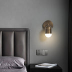 Single-capsule antique brass Clérachic wall light on a grey wall, above a black bedside table and a leather headboard.