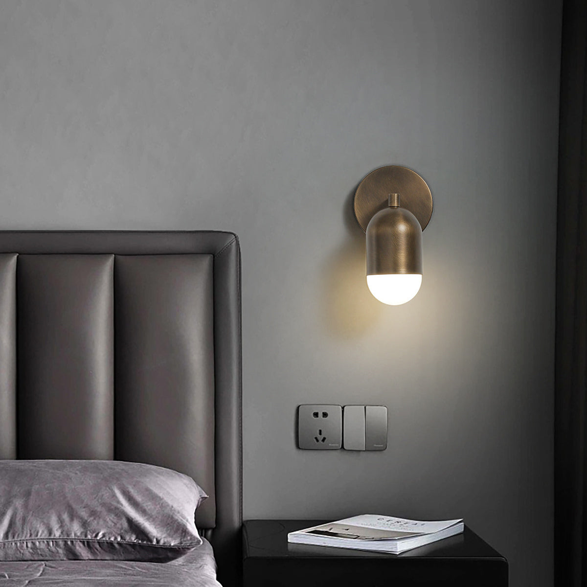 Single-capsule antique brass Clérachic wall light on a grey wall, above a black bedside table and a leather headboard.