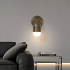 An antique brass Clérachic wall light with a downward capsule shade, above a black leather sofa and a glass side table.