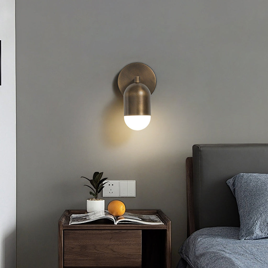 Antique brass Clérachic wall light with a single glowing shade, above a walnut nightstand with a plant and a magazine.