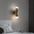 A Clérachic wall light with two brass capsule shades lit top and bottom, above a bed with grey bedding.