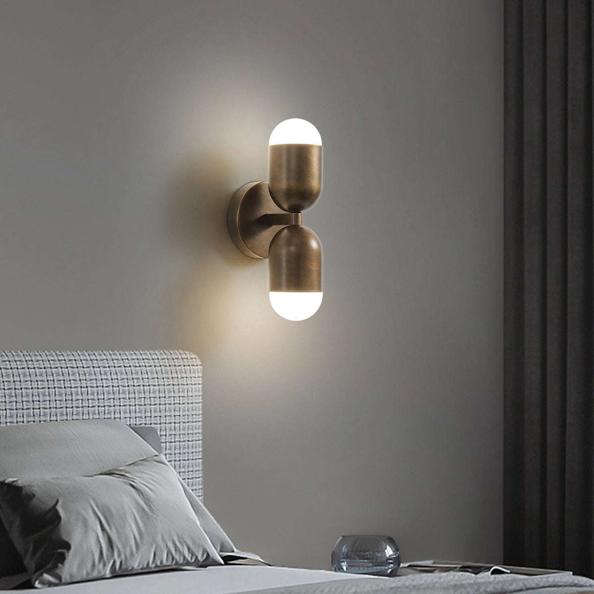 A Clérachic wall light with two brass capsule shades lit top and bottom, above a bed with grey bedding.