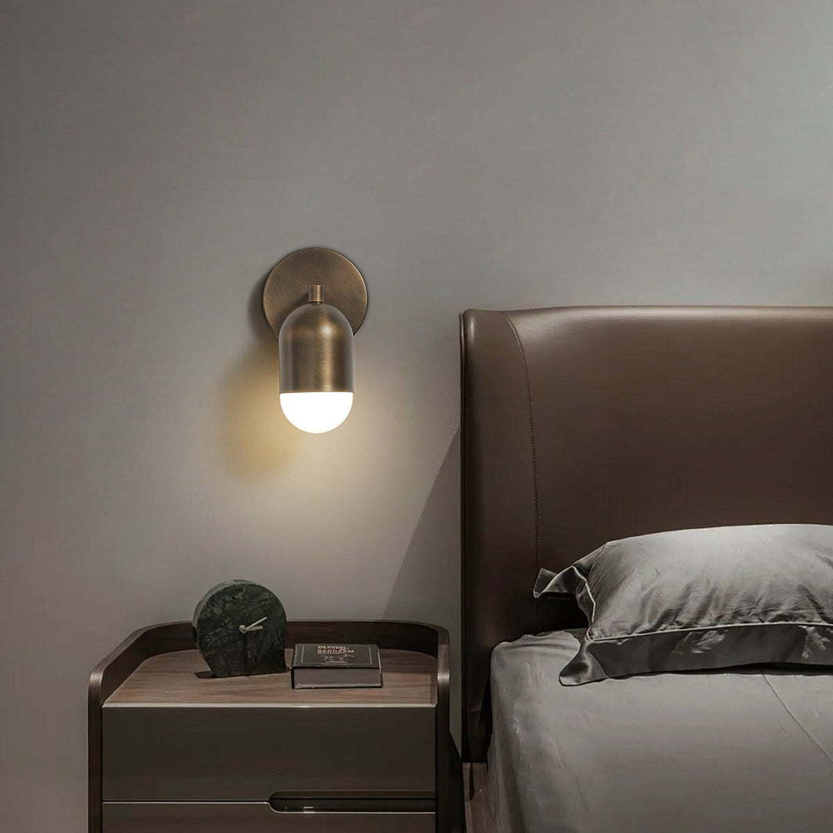 Single antique brass Clérachic wall light glowing above a walnut bedside table and a brown leather headboard.