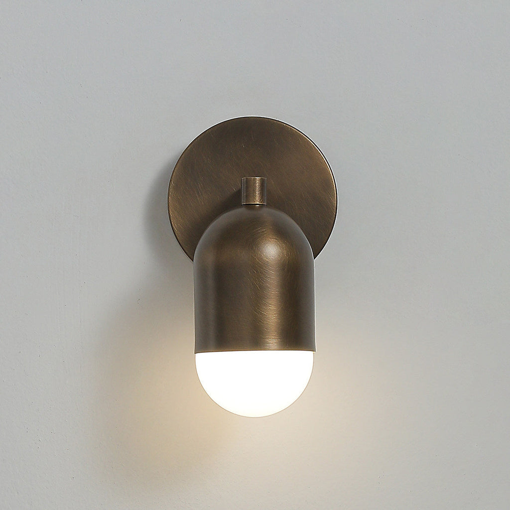Close, front-on view of an antique brass Clérachic wall light with one lit frosted capsule below a round backplate.