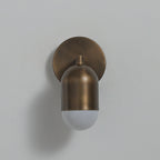 Antique brass Clérachic wall light with its frosted capsule unlit, hanging below a round backplate on a pale grey wall.