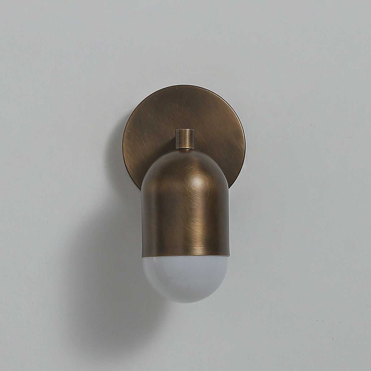 Antique brass Clérachic wall light with its frosted capsule unlit, hanging below a round backplate on a pale grey wall.