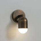 Side view of an antique brass Clérachic wall light with a lit frosted capsule and a round backplate, on a grey wall.