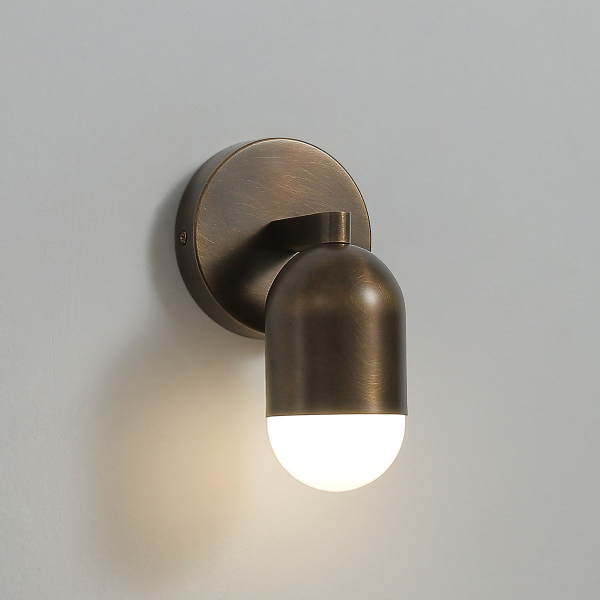 Side view of an antique brass Clérachic wall light with a lit frosted capsule and a round backplate, on a grey wall.