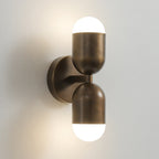 A double-capsule antique brass Clérachic wall light with both frosted ends lit, on a pale wall.