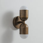 Antique brass Clérachic wall light with two frosted capsule ends, unlit, seen from the side on a grey wall.