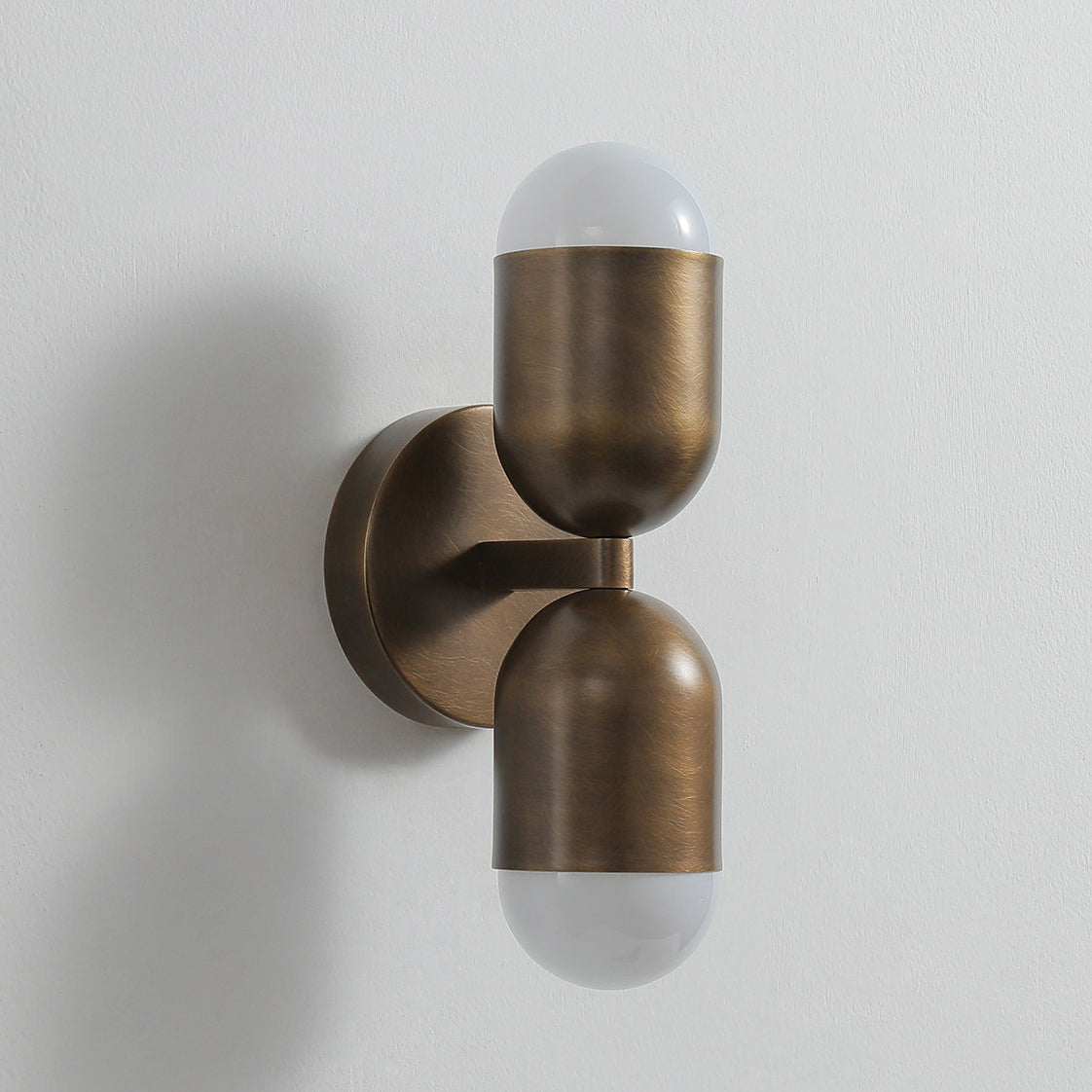 Antique brass Clérachic wall light with two frosted capsule ends, unlit, seen from the side on a grey wall.
