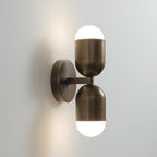 Side view of a two-capsule antique brass Clérachic wall light, both frosted ends glowing, on a pale wall.
