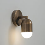 A single-capsule antique brass Clérachic wall light with its frosted end lit, on a pale grey wall.