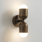 Antique brass Clérachic wall light with two capsule shades, both lit, on a pale grey wall.