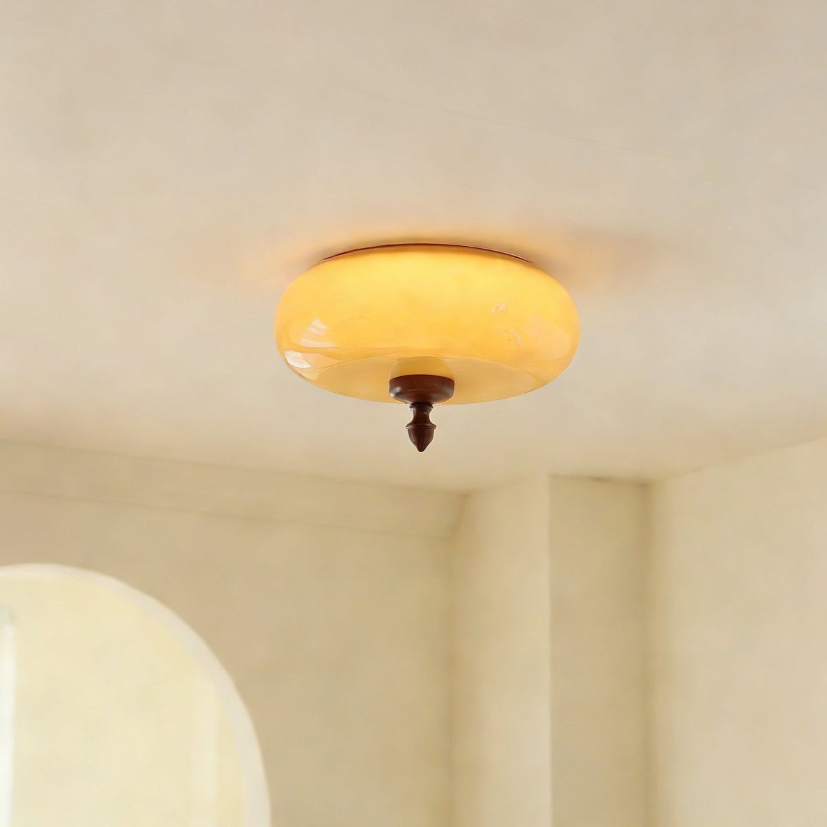 Close-up from below of the glowing amber glass shade and walnut finial of this Clérachic ceiling light against a cream ceiling.