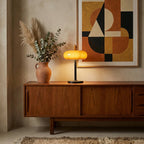 Small Clérachic table lamp glowing on a walnut sideboard, with dried pampas grass in a clay vase and a geometric print behind.