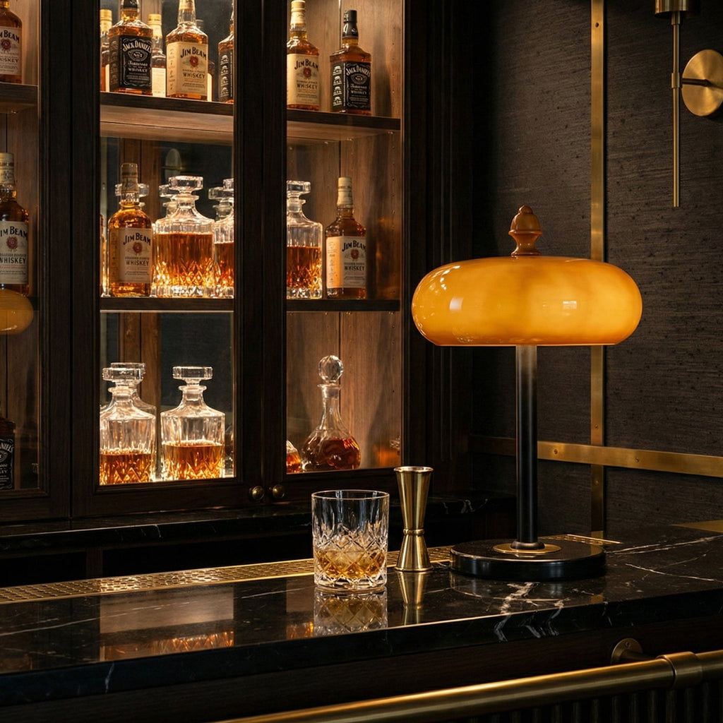 Glowing caramel glass lamp from Clérachic on a black marble bar top, with whisky decanters and a tumbler in the cabinet behind.