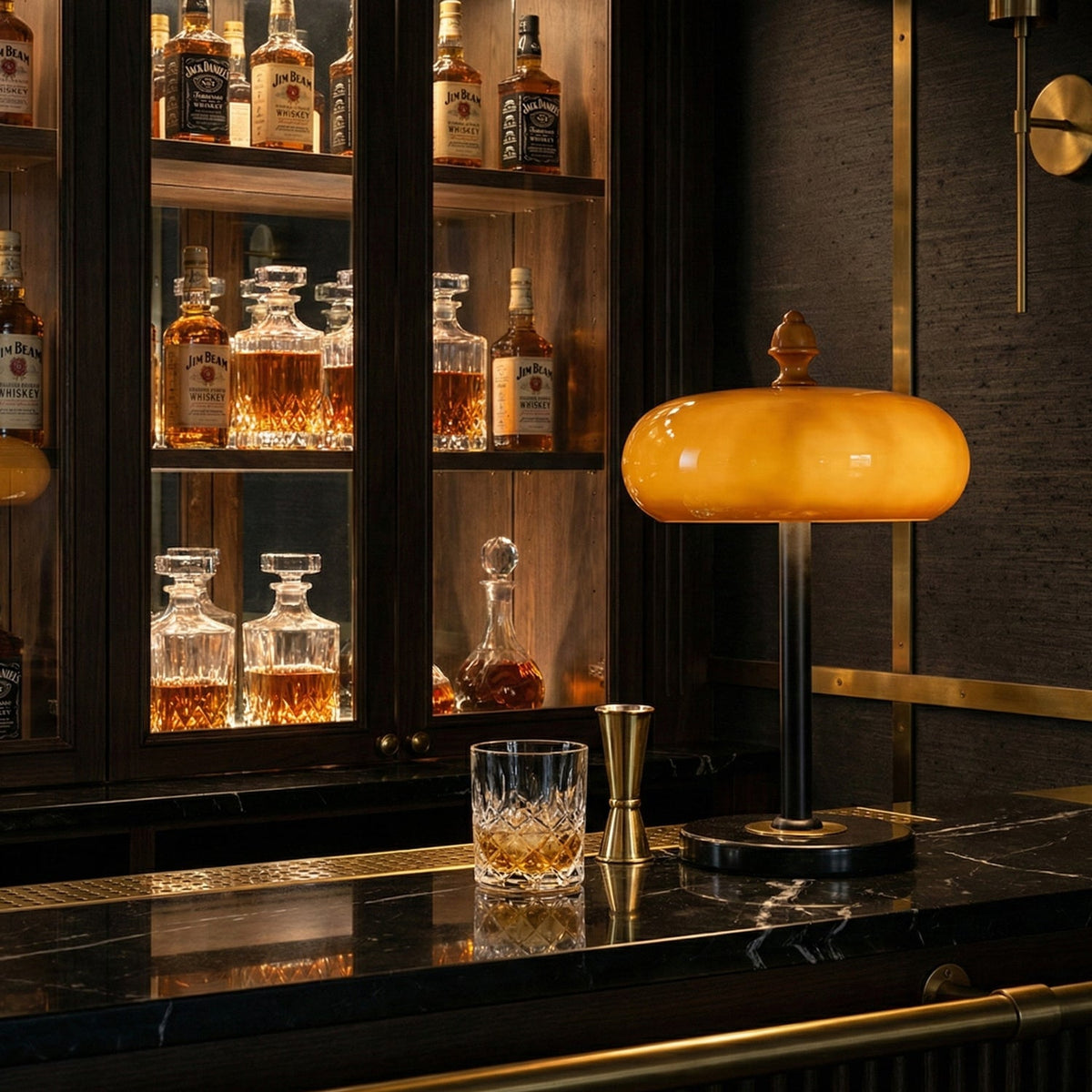 Glowing caramel glass lamp from Clérachic on a black marble bar top, with whisky decanters and a tumbler in the cabinet behind.