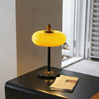 Lit caramel dome lamp from Clérachic on a black side table with a folded newspaper, near a window with open shutters.