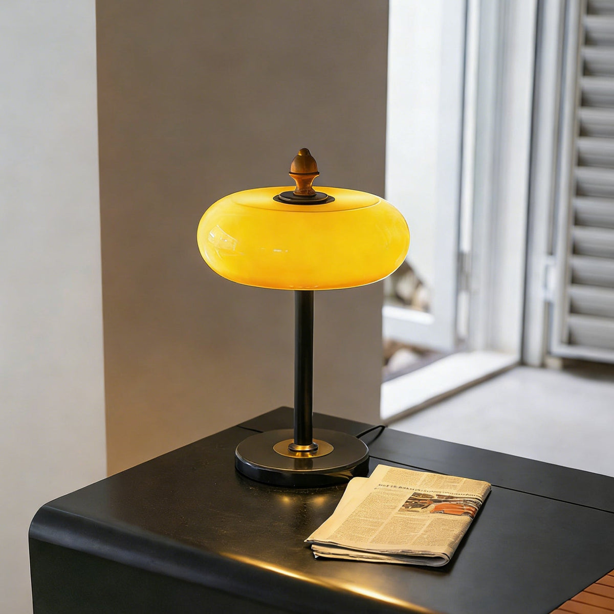 Lit caramel dome lamp from Clérachic on a black side table with a folded newspaper, near a window with open shutters.