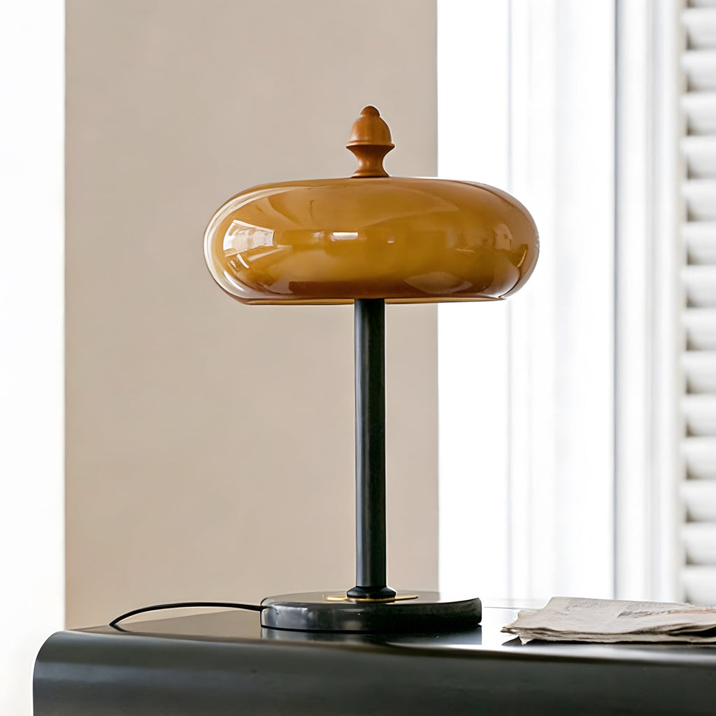 Clérachic table lamp unlit, its smooth caramel glass dome and wooden finial in front of a grey wall and a white shutter.
