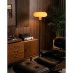 Lit caramel shade of this Clérachic floor lamp against walnut panelling, near a record player, a drawer unit and a black leather lounge chair.