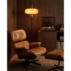 Warm amber light from a Clérachic floor lamp falls across a tan leather lounge chair and a turntable cabinet in a wood-clad room.