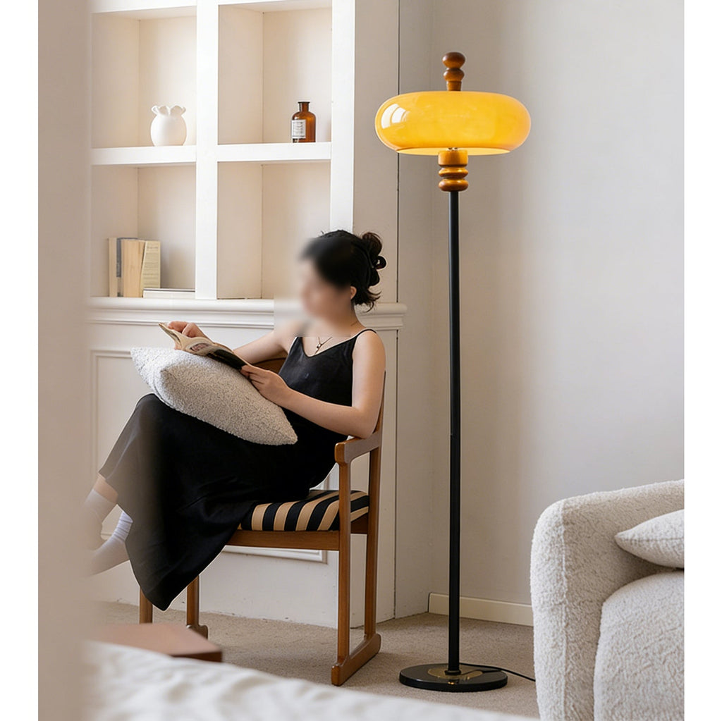 A woman reads in a wooden chair next to this Clérachic floor lamp, its caramel shade lit, in front of white built-in shelves.