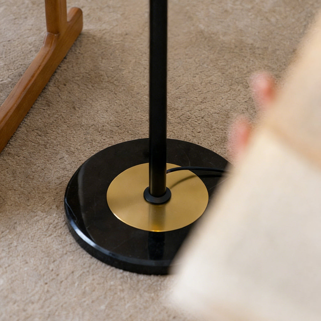 Close view of the round black marble base, brass disc and black stem of this Clérachic floor lamp, standing on a carpet.